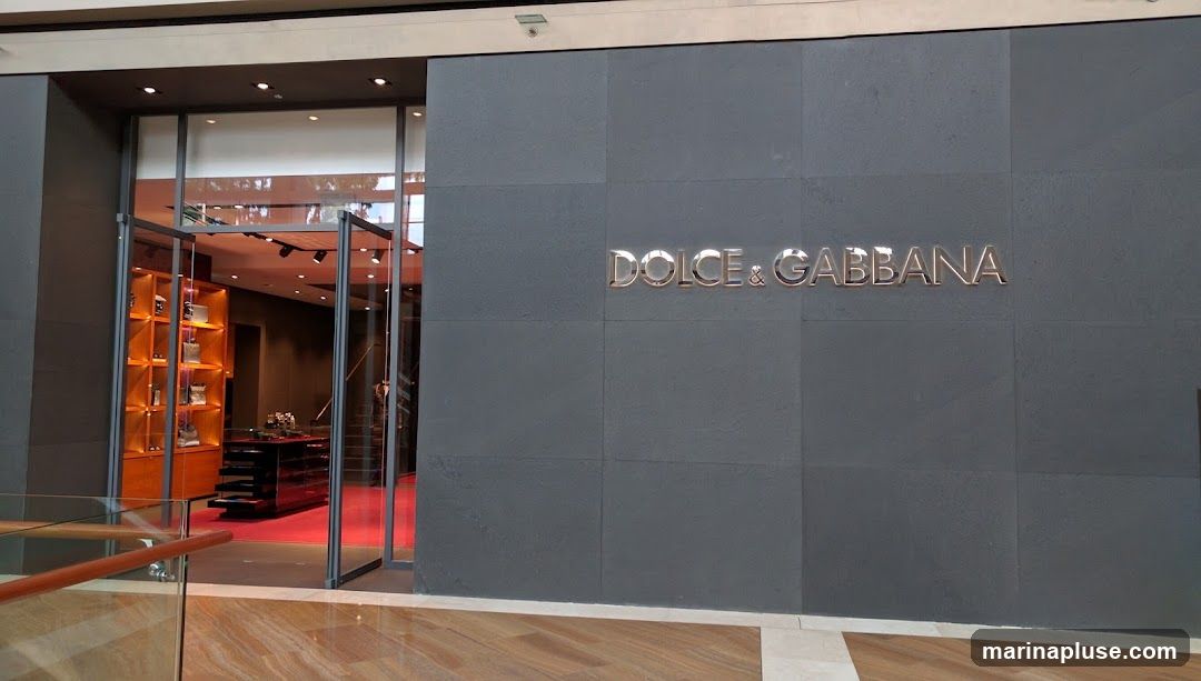 The exterior of a Dolce & Gabbana store. The facade is dark gray with the store's name in large, metallic letters. To the left, glass doors open into the store, revealing shelves stocked with merchandise and a red carpeted floor.