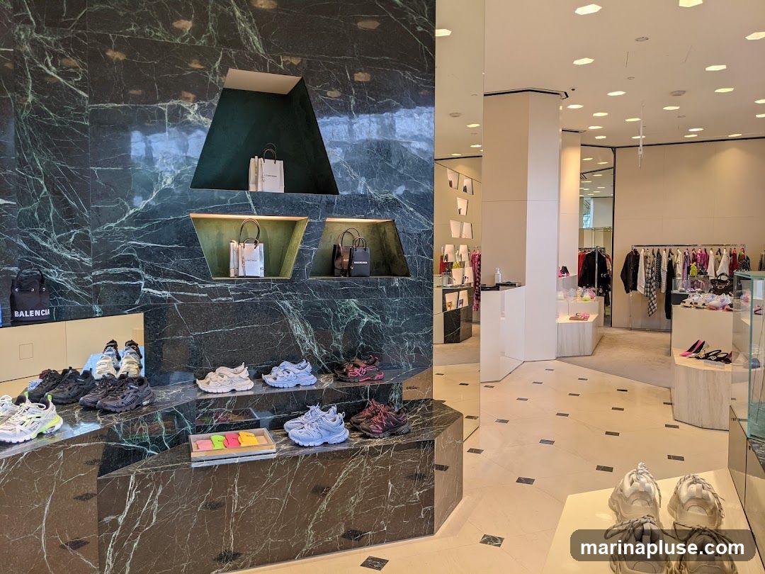 A view of the interior of a Balenciaga store, showcasing a display of sneakers and handbags on marble shelves. In the background, racks of clothing and more footwear are visible, along with display cases. The store has a modern and luxurious aesthetic with marble accents and bright lighting.