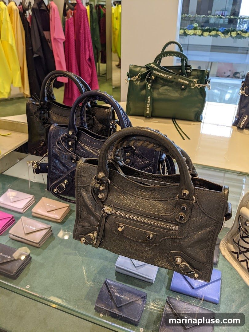 A display of Balenciaga handbags and wallets in various colors, including black, navy, and green. The handbags are on a shelf, and the wallets are arranged on a glass surface. In the background, racks of colorful clothing are visible.