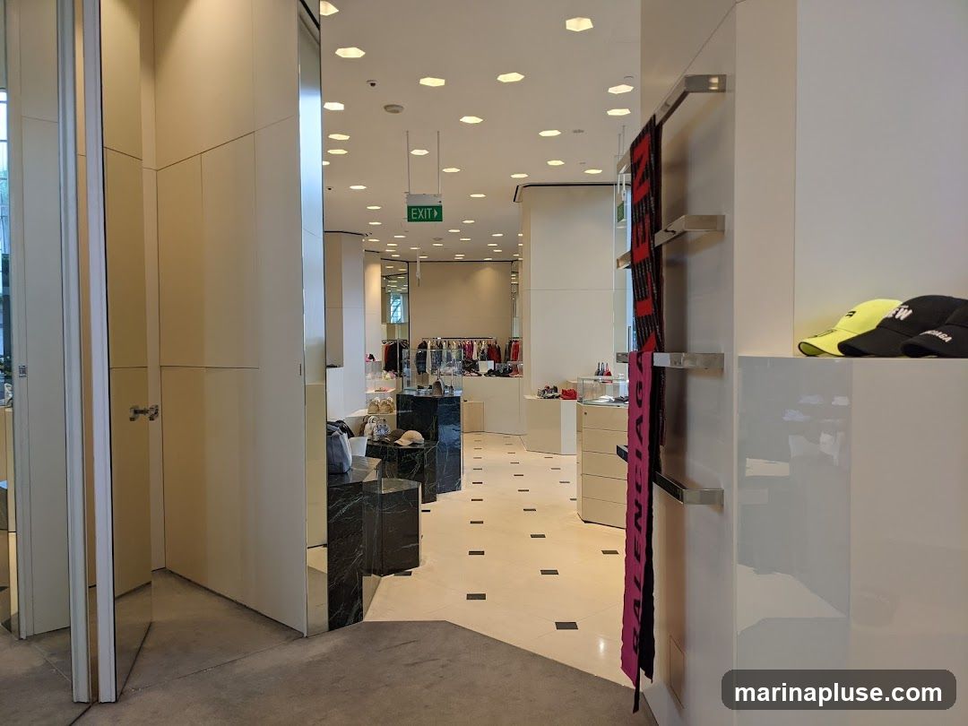 The image shows the interior of a retail store, likely a clothing boutique. In the foreground, a white shelf displays two baseball caps, one yellow and one black, with visible branding. To the left, a display rack holds several scarves, one pink and one black with red lettering, also featuring branding. Further into the store, clothing racks with various garments are visible, along with display cases and marble pedestals. An "EXIT" sign is illuminated in green overhead. The overall aesthetic is modern and minimalist, with polished floors and bright lighting.