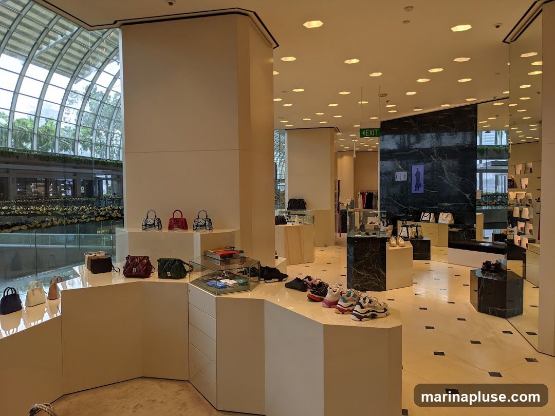 A well-lit interior of a luxury retail store showcasing a collection of designer handbags and sneakers. The display features minimalist white shelving and geometric pedestals, with handbags arranged on tiered shelves and sneakers displayed on a lower counter. The store's interior is modern and elegant, with marble accents and a mirrored wall reflecting the merchandise. In the background, racks of clothing and additional display cases are visible. The overall ambiance suggests a high-end shopping experience.