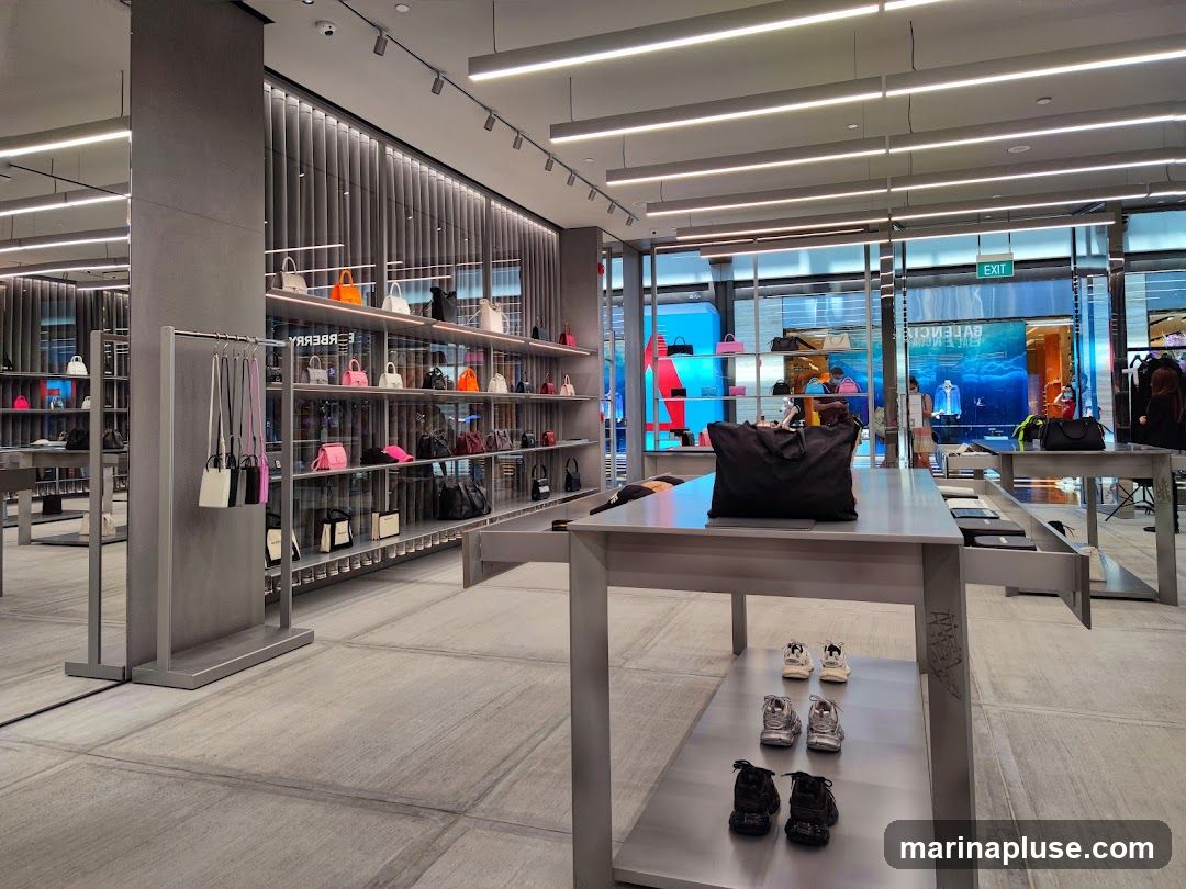 The interior of a modern, upscale retail store featuring a wide selection of designer handbags displayed on shelves and racks. In the foreground, tables showcase various styles of sneakers. The store has a minimalist design with grey walls, polished floors, and bright overhead lighting. Through the glass facade, an 'EXIT' sign is visible, along with glimpses of the street outside.