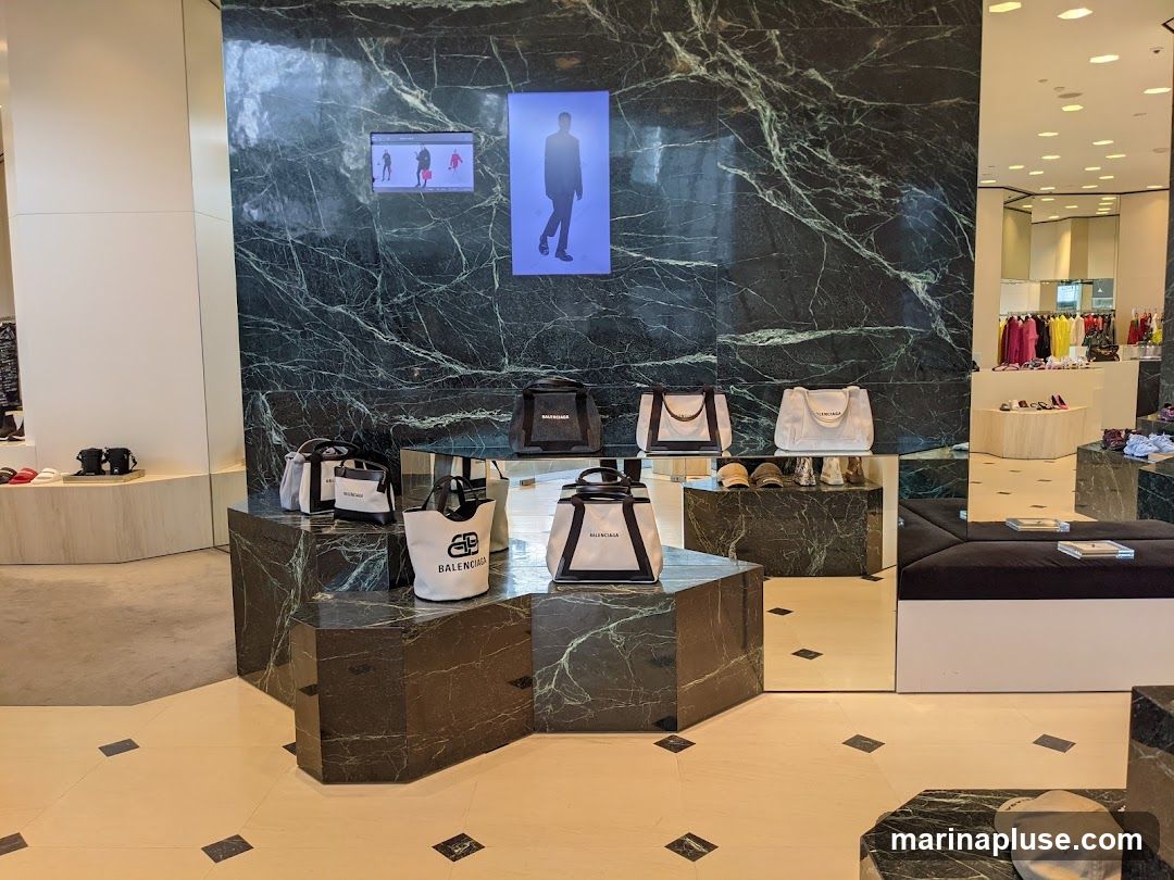 A retail store interior with a display of Balenciaga handbags on a marble-patterned wall and hexagonal pedestals. Digital screens show models. In the background, racks of clothing and shoes are visible.