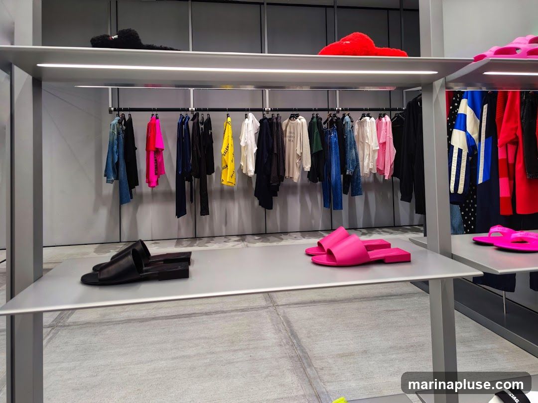 A retail display featuring various clothing items and footwear. On the top shelf, there are fuzzy slippers in black and red. Below, a rack of colorful clothing, including t-shirts, jackets, and pants, is displayed. On the lower shelves, there are black slides and bright pink slides. Further to the right, more clothing is visible, including a blue and white striped jacket and red garments. The store has a minimalist aesthetic with grey walls and shelving.