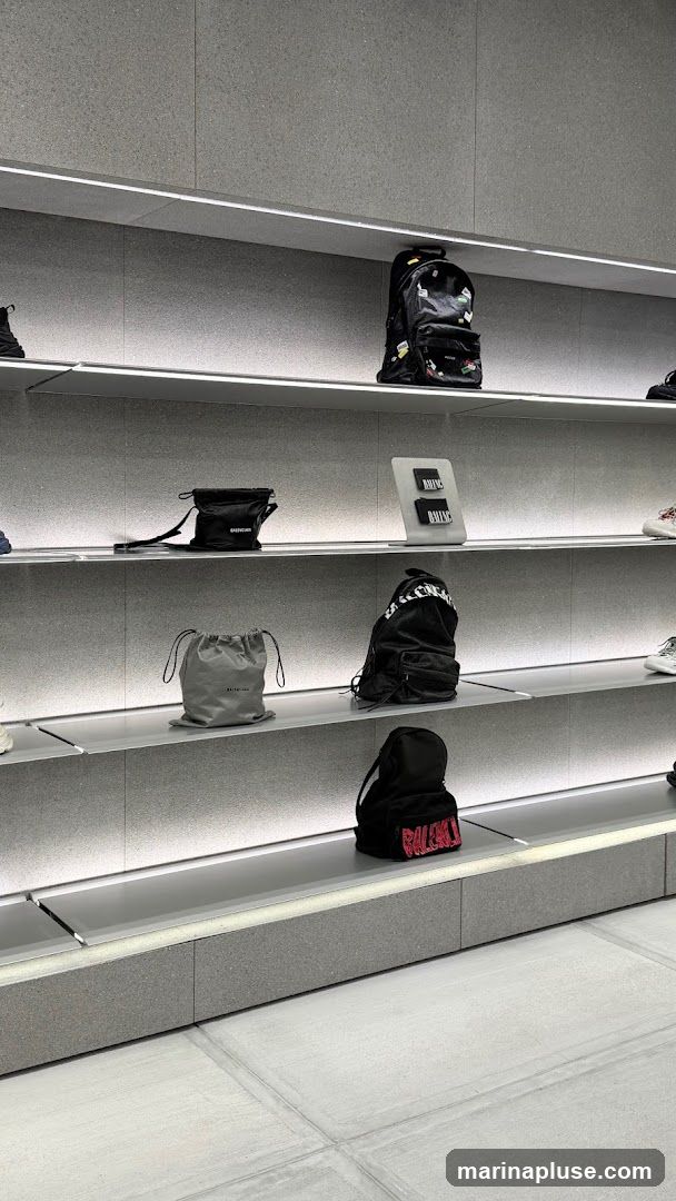 Shelves in a store display various bags and accessories. On the top shelf, a black backpack adorned with patches is visible. Below it, a black crossbody bag with a strap and a grey drawstring bag are displayed. Further down, another black backpack with white lettering and a smaller black backpack with red lettering are shown. A display stand with two black cards featuring white text is also present.