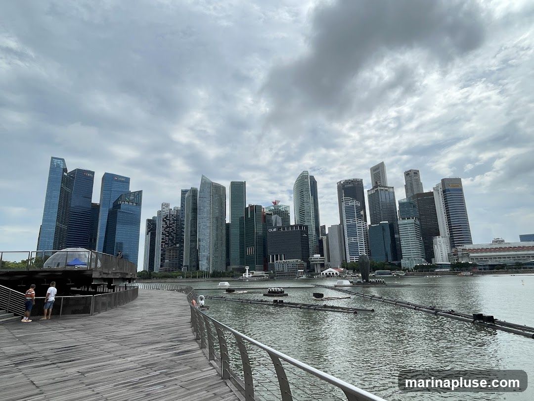 A wide shot of the Singapore skyline with its modern skyscrapers on a cloudy day. A wooden walkway with a metal railing curves into the foreground, leading towards the water. Two people are standing on the walkway, looking out at the view. In the water, there are some floating structures and buoys.