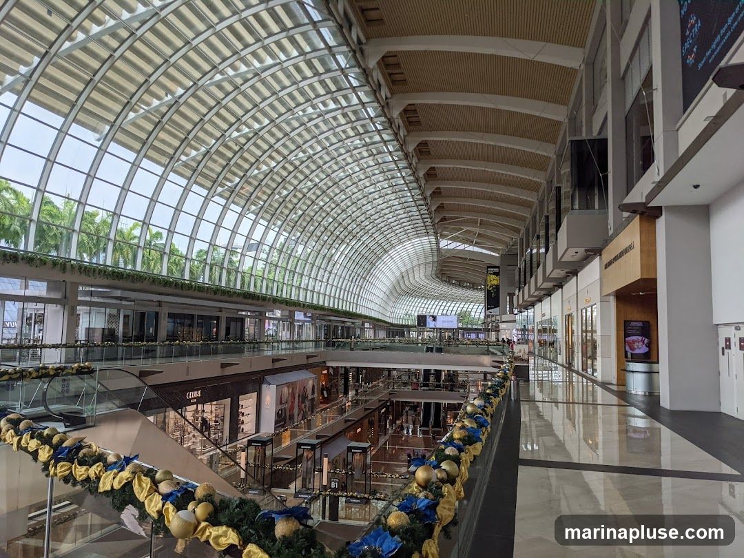 A wide-angle view of the interior of a large, modern shopping mall. The architecture features a distinctive curved glass roof that allows natural light to flood the space. Multiple levels of shops are visible, with escalators and walkways connecting them. Decorative garlands with gold and blue accents are draped along the railings, suggesting a festive season. The mall appears spacious and well-lit, with a clean and contemporary aesthetic.