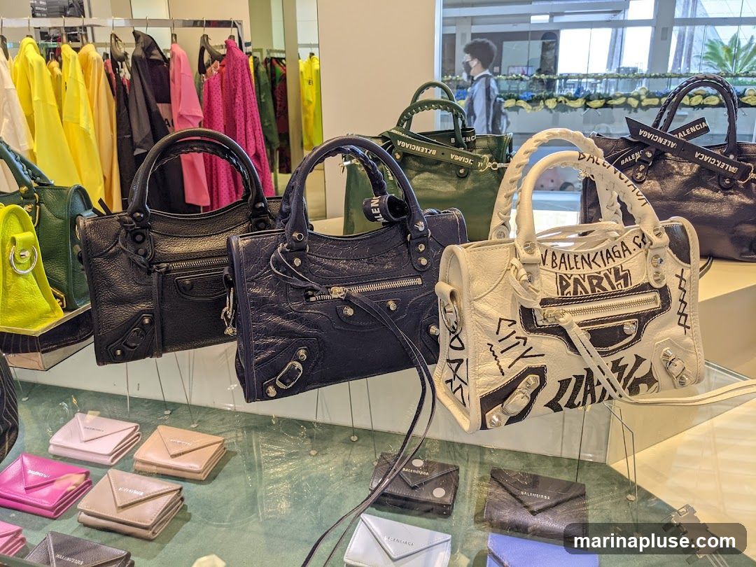 A display of various designer handbags and small leather goods, including wallets and cardholders, in a retail setting. The handbags are in different colors such as black, navy, green, and white, with the white bag featuring graffiti-style artwork. The smaller items are neatly arranged in rows on a glass surface. In the background, racks of colorful clothing are visible, suggesting a fashion boutique.