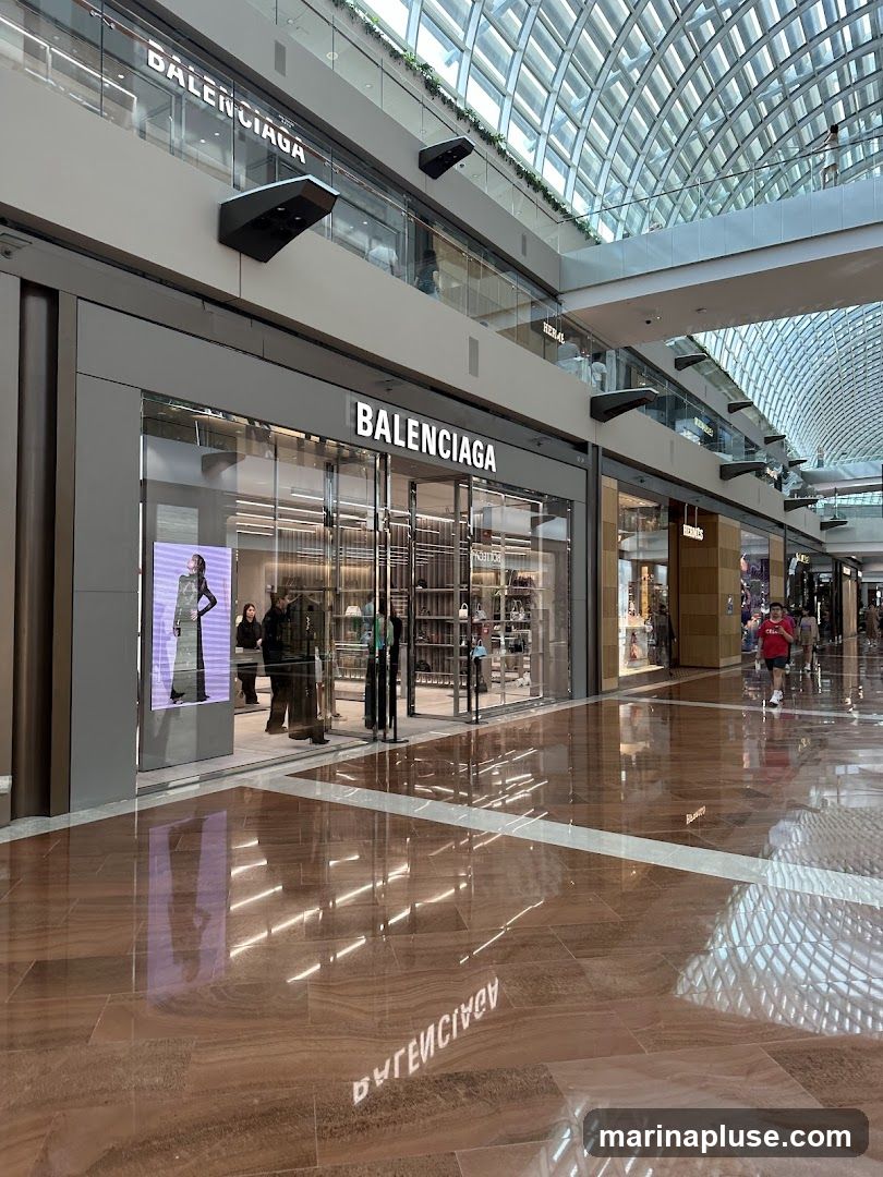 The exterior of a Balenciaga store located in a modern indoor shopping mall. The store has a large glass facade with the Balenciaga logo prominently displayed in white lettering above the entrance. Inside, merchandise is visible on display shelves. The floor is highly polished, reflecting the store and the mall's architecture.