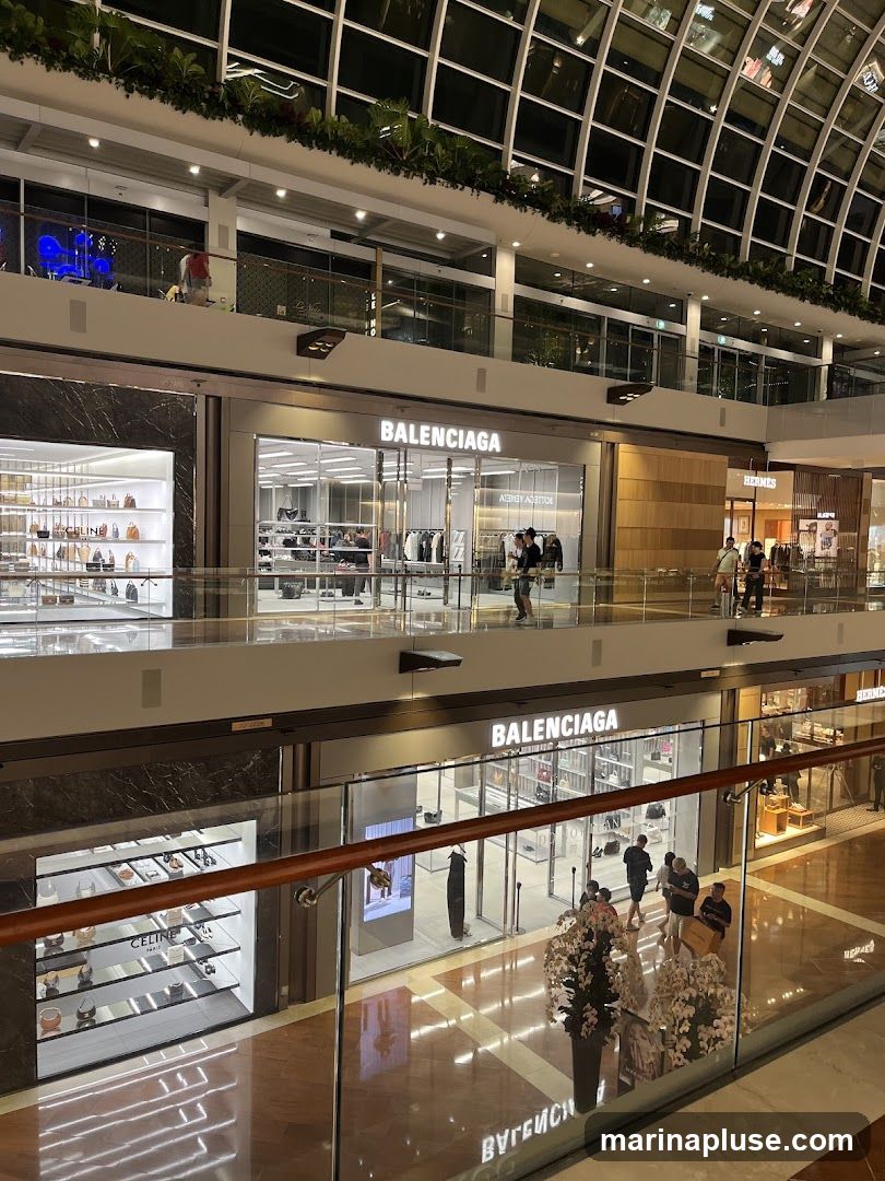 A view of multiple luxury brand stores on different levels of a shopping mall. The stores visible include Balenciaga, Celine, and Hermes. The architecture of the mall features a large, arched glass ceiling with internal lighting and greenery. People are seen browsing inside and walking between the shops.