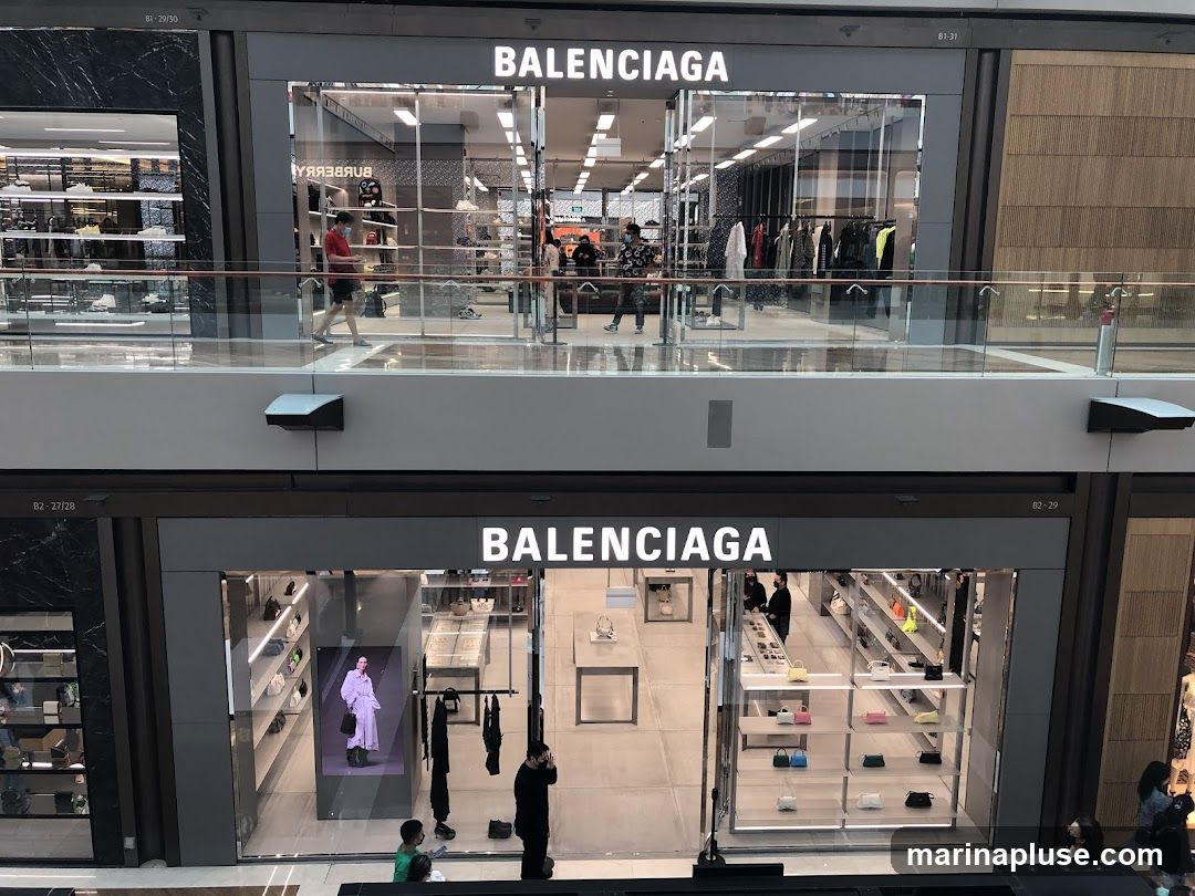 A high-angle view of two Balenciaga storefronts on different levels of a shopping mall. The upper storefront displays clothing on racks and mannequins, while the lower storefront showcases handbags on shelves and a digital display featuring a model. Both storefronts have prominent "BALENCIAGA" signage.