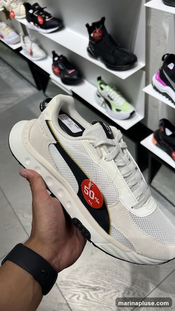 A hand holds a white and beige Puma sneaker with black accents. A red oval tag with "50%" and the Puma logo is attached to the side of the shoe. The sneaker is displayed in a store with other shoes on shelves in the background.