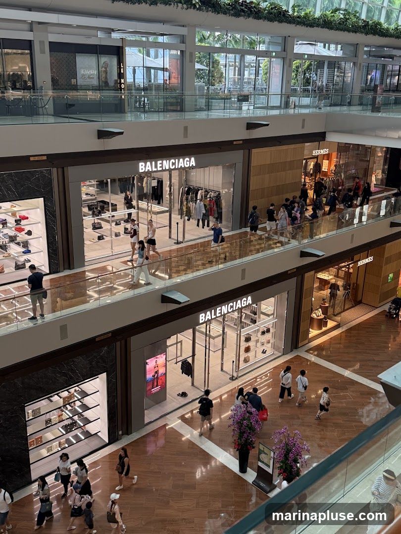 An overhead view of two Balenciaga stores located on different levels of a modern shopping mall. The stores feature large glass windows displaying clothing and handbags. People are seen browsing inside and walking in the mall corridors. Other luxury brands like Hermes are also visible.