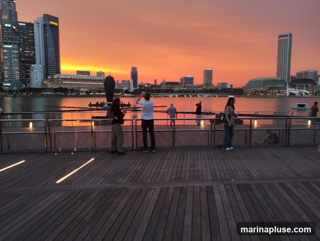 A group of people are standing on a wooden boardwalk, watching the sunset over a city skyline. The sky is a vibrant orange and yellow, reflecting on the water. The city features modern skyscrapers and a domed building.