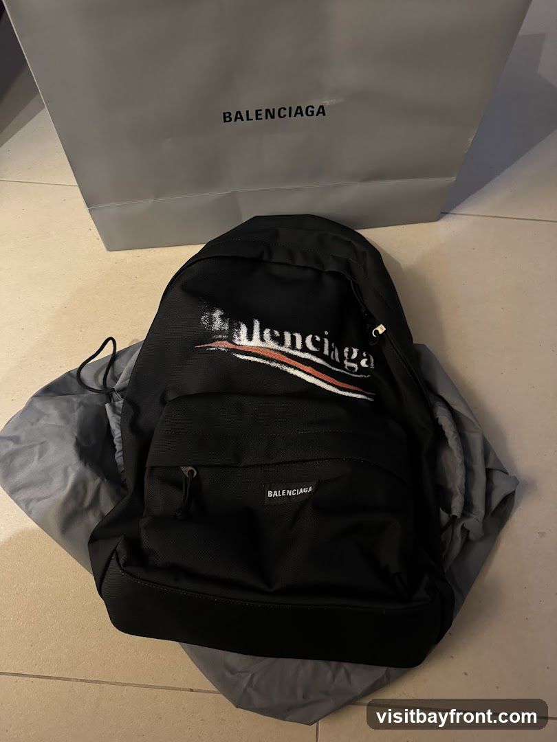 A black Balenciaga backpack with a distressed logo print and a smaller Balenciaga logo on the front pocket is displayed. The backpack is placed on a grey dust bag. Behind the backpack is a grey Balenciaga shopping bag with the brand name printed in black.