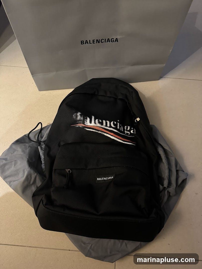 A black Balenciaga backpack with a distressed logo print and a smaller Balenciaga logo on the front pocket is displayed. The backpack is placed on a grey dust bag. Behind the backpack is a grey Balenciaga shopping bag with the brand name printed in black.