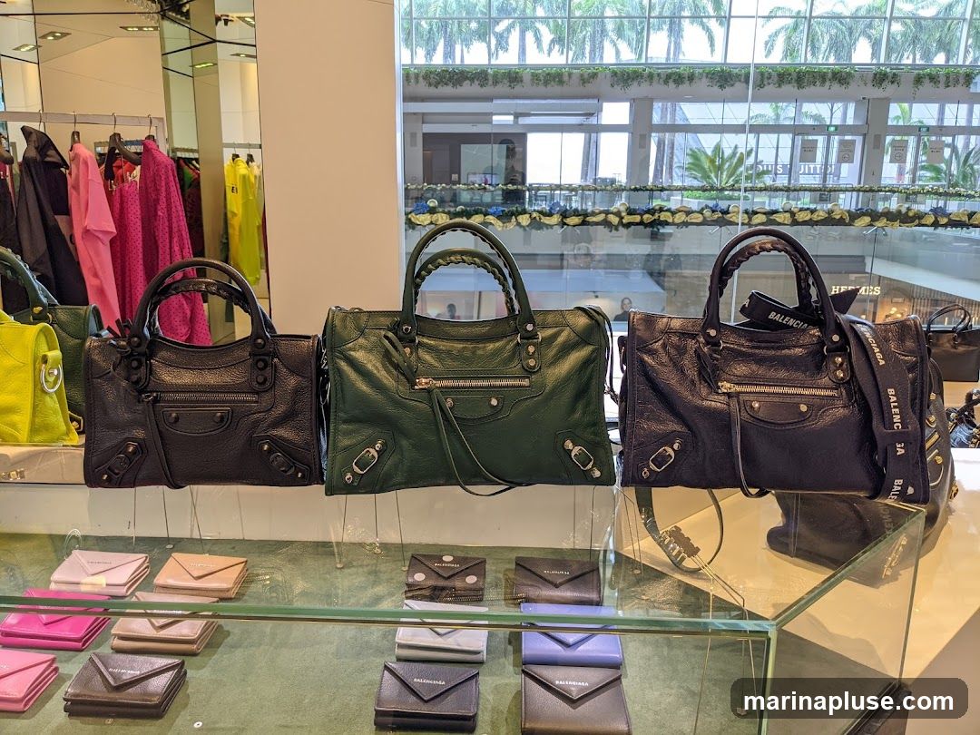A display of designer handbags and wallets in a store. Three handbags are prominently featured on a shelf, with a selection of smaller wallets and cardholders arranged below. The handbags are in black and dark green leather, featuring the brand's signature hardware and braided handles. The wallets are in various colors including pink, beige, grey, and purple. In the background, racks of colorful clothing are visible, along with reflections in mirrors and a view of a shopping mall interior through a large window.