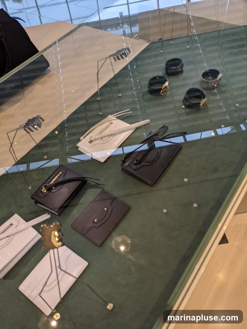 A glass display case shows various luxury accessories, including black and white clutches, wallets, and several black and grey bracelets with gold accents. The items are arranged on a dark green velvet surface. The reflection of the ceiling and windows is visible on the glass.