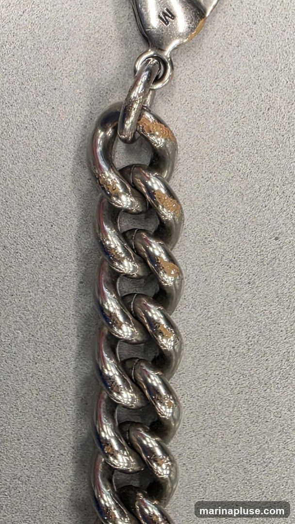 A close-up shot of a thick, silver-colored chain with visible wear and some tarnishing on the links. A small tag with the letter 'M' is attached to the top link. The chain is resting on a textured gray surface.