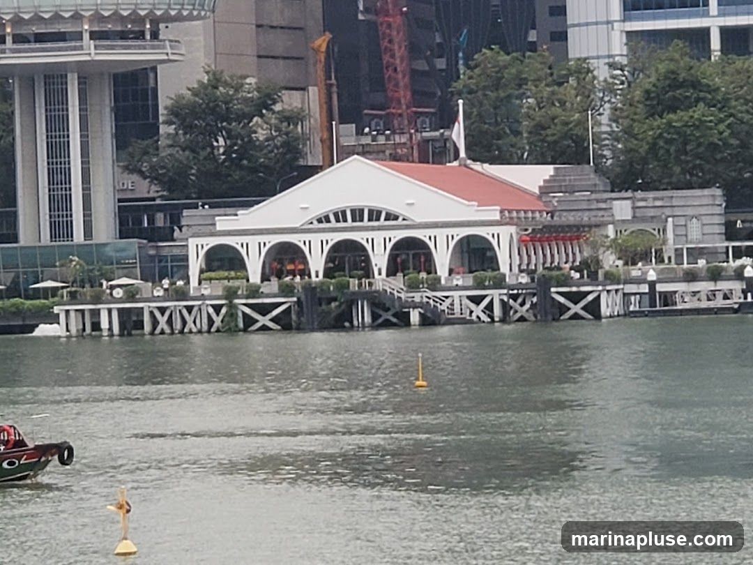 A white building with a red roof and arched openings stands on a pier over the water. In the background, modern high-rise buildings and trees are visible. A boat is in the foreground on the water.