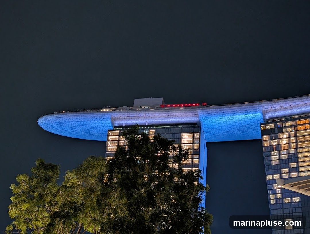 A nighttime view of the Marina Bay Sands hotel in Singapore, with its iconic rooftop infinity pool illuminated in blue. The hotel's distinctive architecture is visible, with lights on in many of the rooms. Trees in the foreground partially obscure the view.