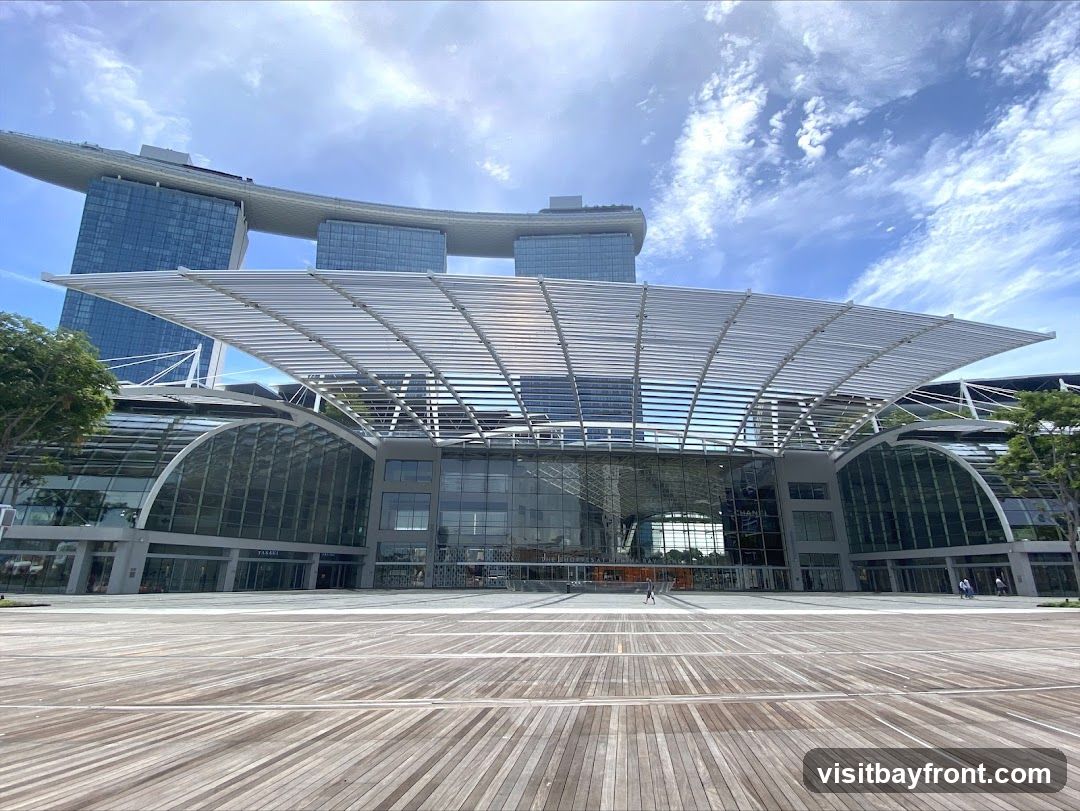 A wide shot of the exterior of the Marina Bay Sands Convention Centre on a sunny day. The building features a large, modern glass facade with a distinctive arched roof structure. A wooden boardwalk stretches out in front of the entrance. The iconic Marina Bay Sands hotel towers in the background.