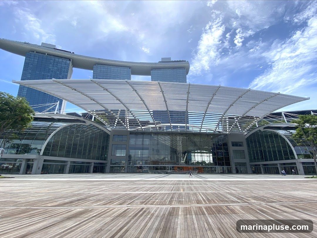 A wide shot of the exterior of the Marina Bay Sands Convention Centre on a sunny day. The building features a large, modern glass facade with a distinctive arched roof structure. A wooden boardwalk stretches out in front of the entrance. The iconic Marina Bay Sands hotel towers in the background.