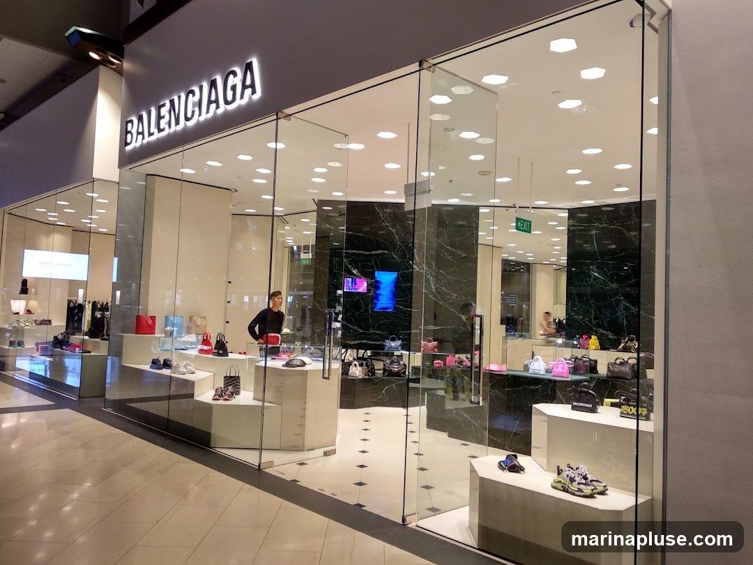 The exterior of a Balenciaga store with large glass windows and doors. The Balenciaga logo is prominently displayed in illuminated white lettering above the entrance. Inside, various handbags and shoes are displayed on tiered shelves and pedestals. A store employee can be seen standing near the entrance.