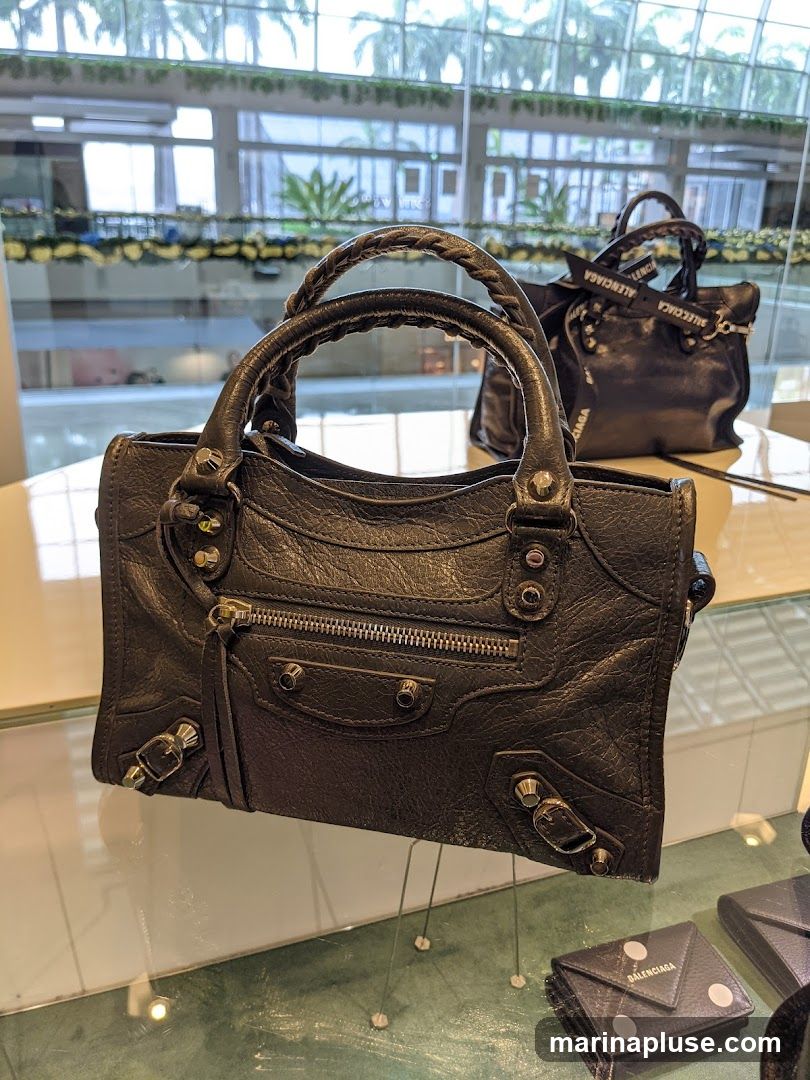 A close-up shot of a black Balenciaga handbag with braided handles and silver hardware. The bag is displayed on a glass surface, with another similar handbag visible in the background. Below the handbag, a small black Balenciaga wallet is also displayed. The background shows a blurred view of a modern interior with large windows and greenery.