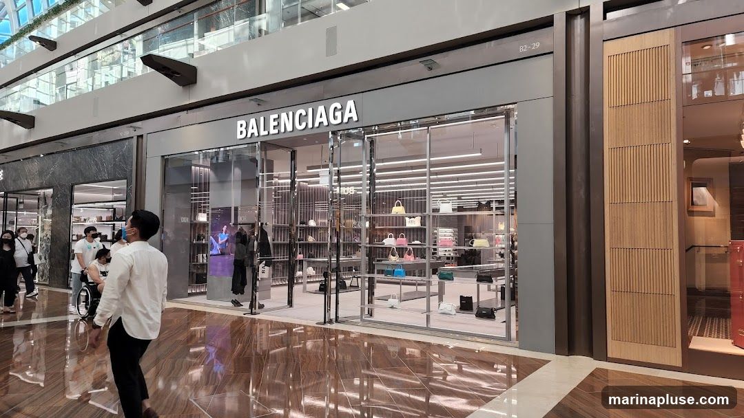The exterior of a Balenciaga store with its name prominently displayed above the entrance. The store features large glass windows showcasing handbags and other merchandise on display shelves. People are seen walking in the mall outside the store.