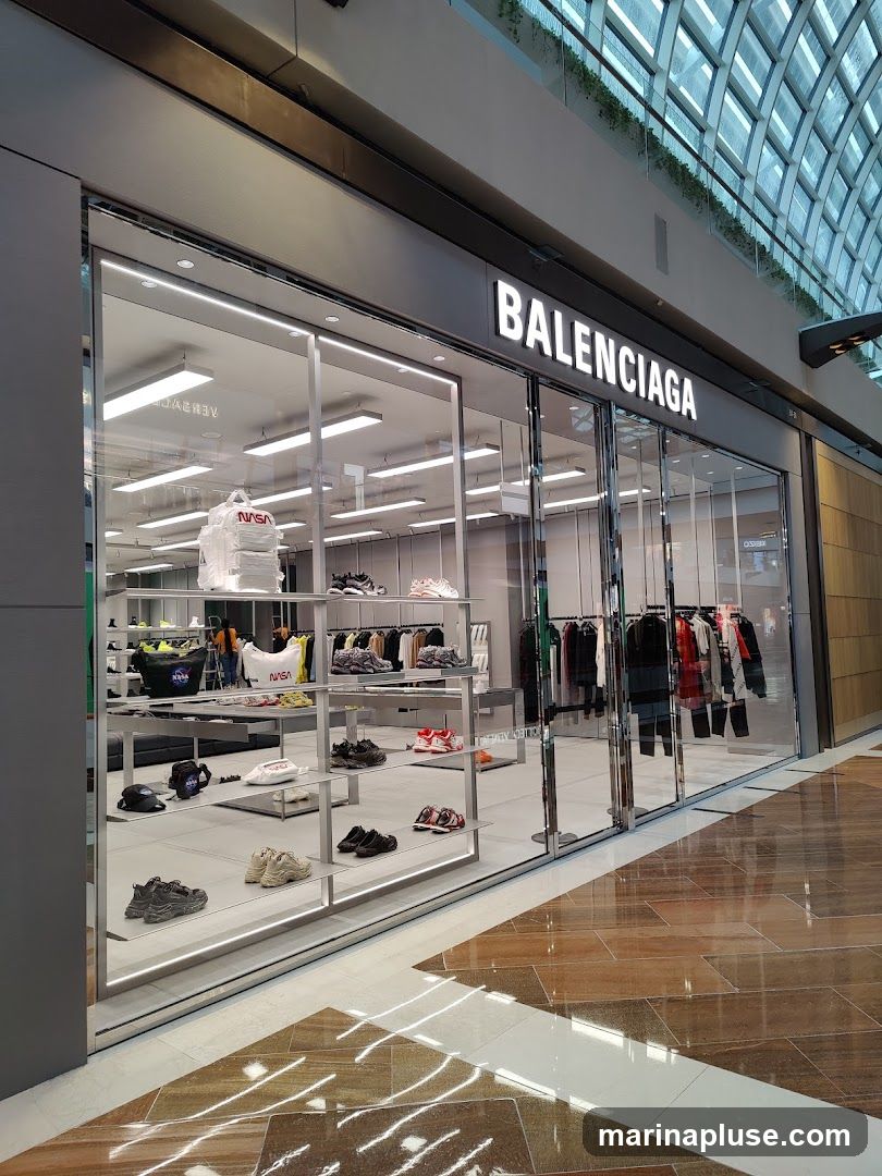 The exterior of a Balenciaga store with large glass windows displaying clothing and shoes. The store's name is prominently displayed in white lettering above the entrance. Inside, shelves are stocked with various footwear and apparel, including items with NASA branding.