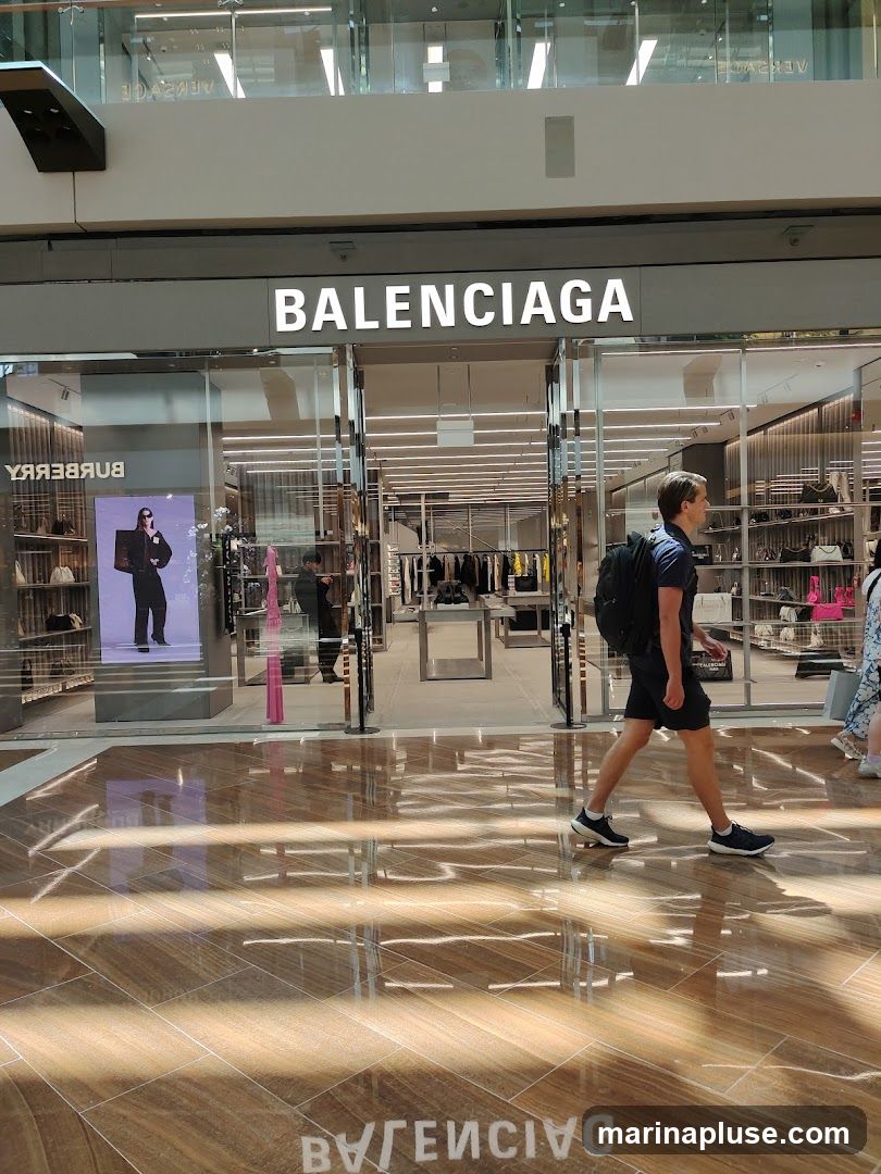 The exterior of a Balenciaga store with the brand name prominently displayed above the entrance. Through the glass doors, racks of clothing and shelves displaying merchandise are visible. A digital display on the left shows a model wearing a black outfit. People are walking past the storefront in a well-lit indoor shopping area with a polished wooden floor that reflects the store's signage.