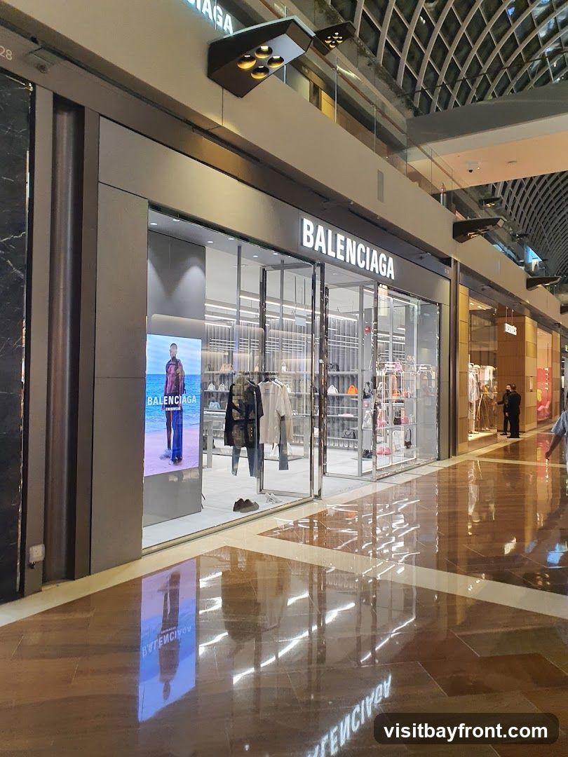 The exterior of a Balenciaga store with its name prominently displayed above the entrance. The store features large glass windows showcasing clothing and accessories. A digital display on the left shows a model. The polished floor reflects the store's interior and signage.