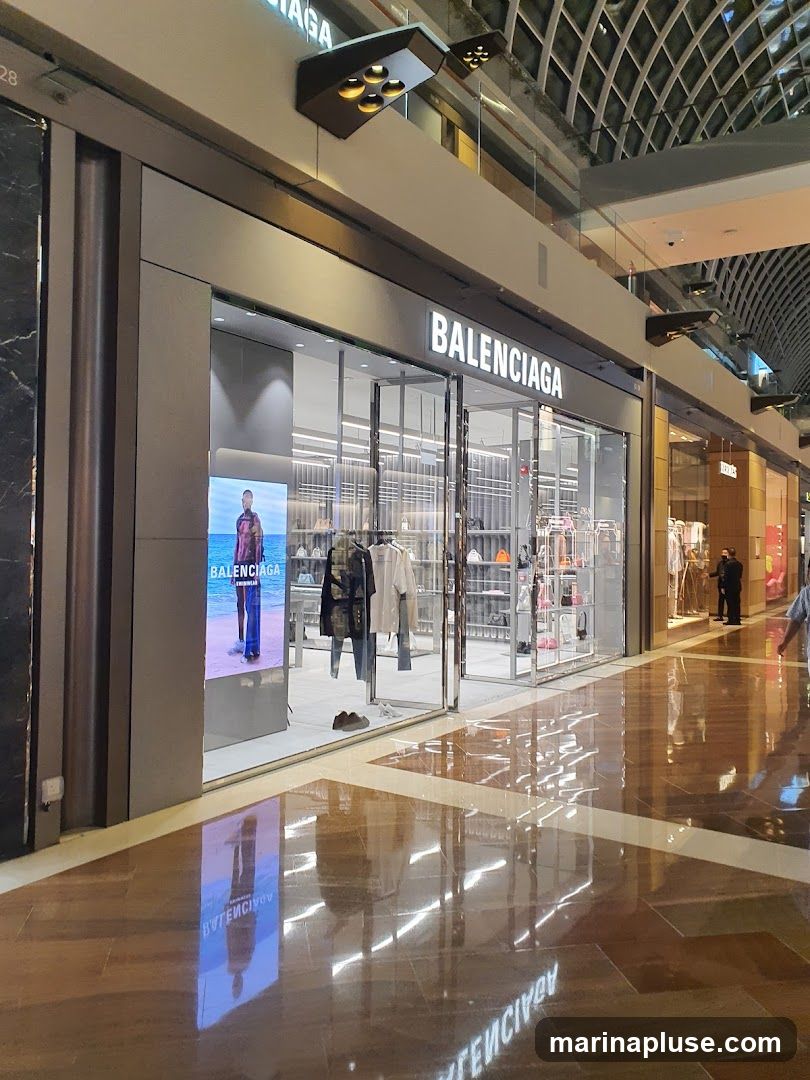 The exterior of a Balenciaga store with its name prominently displayed above the entrance. The store features large glass windows showcasing clothing and accessories. A digital display on the left shows a model. The polished floor reflects the store's interior and signage.