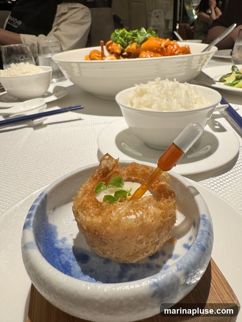 A close-up view of a crispy, hollow appetizer served in a blue and white patterned bowl. A dropper filled with orange sauce is inserted into the appetizer, with a few green herbs on top. In the background, bowls of white rice and a large dish of food in a red sauce are visible, suggesting a meal setting.