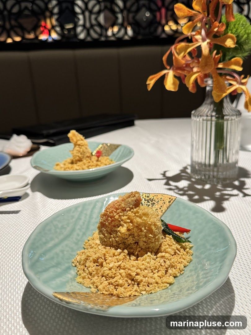 A close-up shot of a dish featuring a single piece of crispy fried shrimp served on a bed of crumbled, golden-brown crumbs. The shrimp is coated in a crunchy batter and garnished with a sliver of red chili and a green leaf. The dish is presented on a light blue, textured plate with gold accents. In the background, another similar dish is visible, along with a vase of orange orchids and a dimly lit, ornate wall decoration. The table is covered with a white tablecloth.