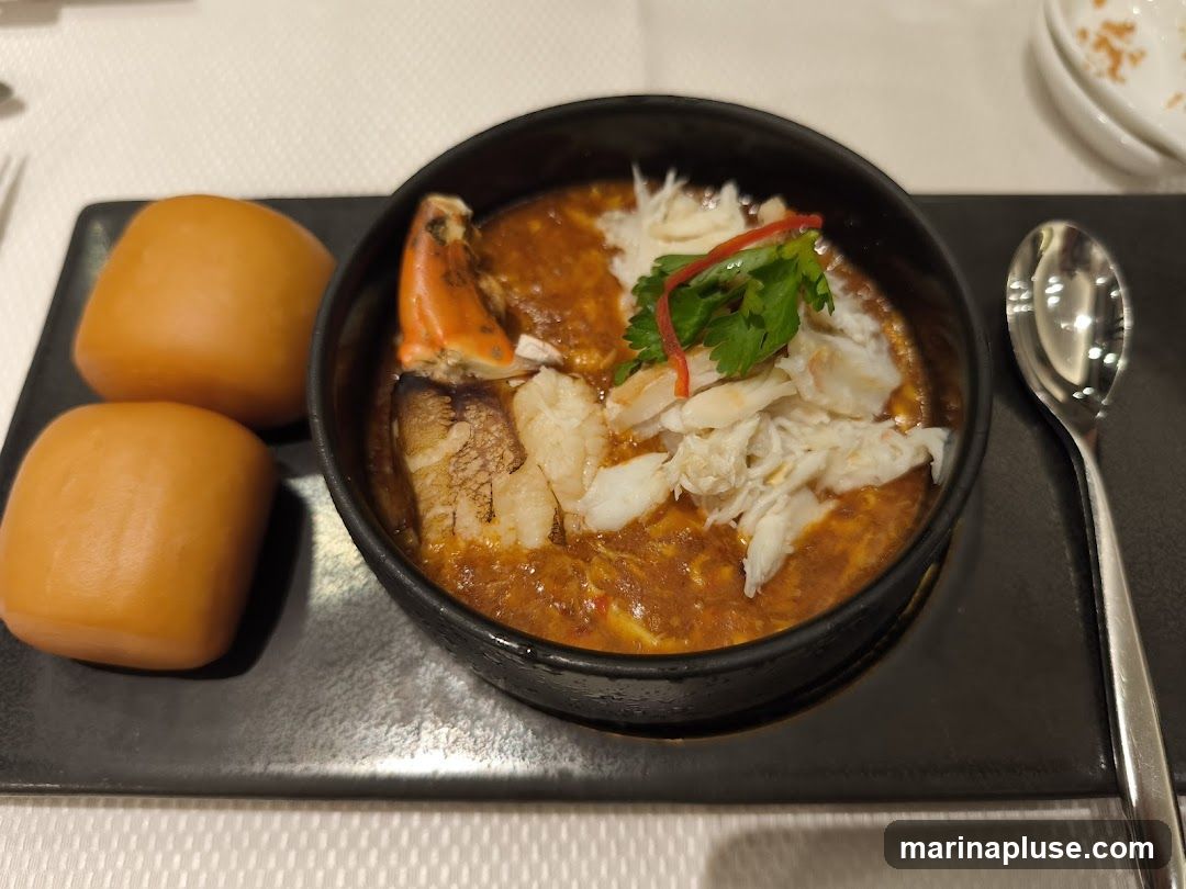 A close-up shot of a crab dish served in a black bowl, accompanied by two steamed buns on a black plate. The crab meat is visible, along with a crab claw and a rich, reddish-brown sauce. The dish is garnished with fresh cilantro and a sliver of red chili. A spoon rests on the right side of the plate.