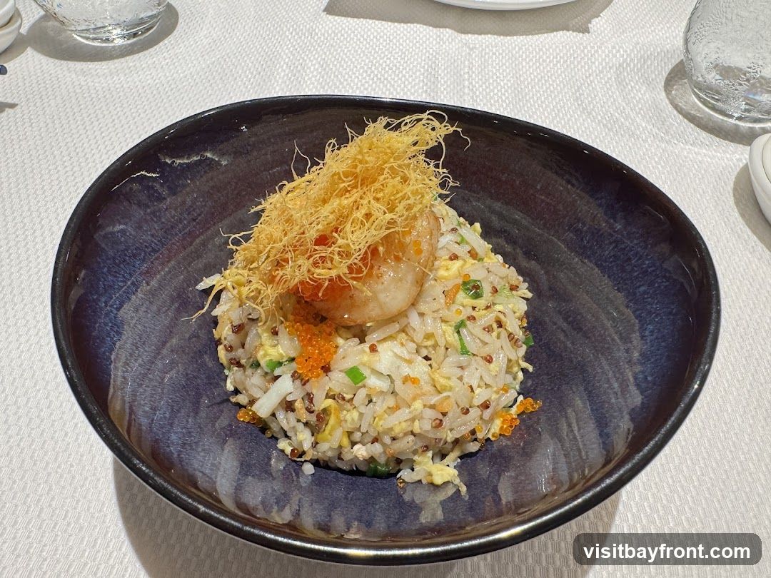 A close-up shot of a bowl of fried rice topped with a single shrimp and crispy fried noodles. The rice is mixed with egg, green onions, and small orange roe. The bowl is dark blue with a glossy finish.