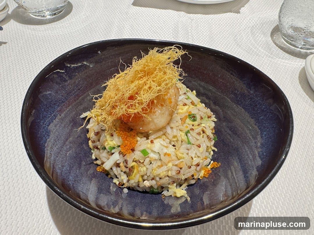 A close-up shot of a bowl of fried rice topped with a single shrimp and crispy fried noodles. The rice is mixed with egg, green onions, and small orange roe. The bowl is dark blue with a glossy finish.