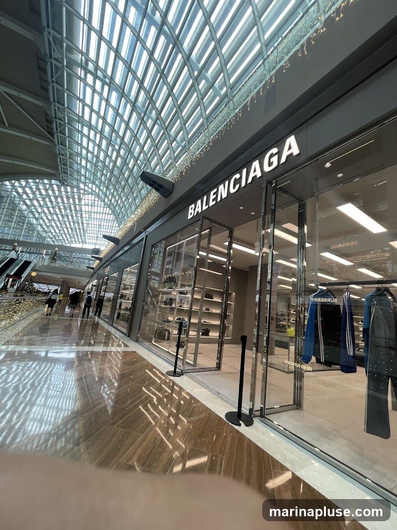 The exterior of a Balenciaga store with its name prominently displayed above the entrance. The store features large glass windows showcasing merchandise, including clothing on racks. The interior is visible, with shelves displaying bags and accessories. The storefront is located within a modern shopping mall with a distinctive arched glass ceiling and reflective flooring.