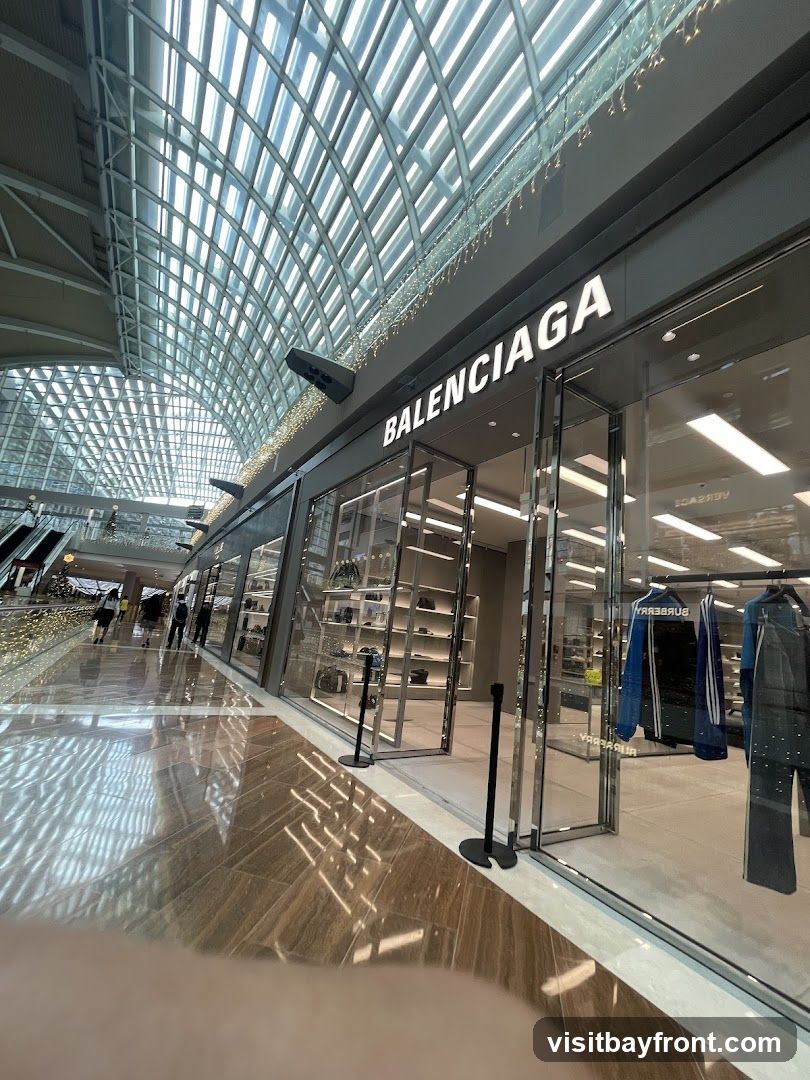 The exterior of a Balenciaga store with its name prominently displayed above the entrance. The store features large glass windows showcasing merchandise, including clothing on racks. The interior is visible, with shelves displaying bags and accessories. The storefront is located within a modern shopping mall with a distinctive arched glass ceiling and reflective flooring.