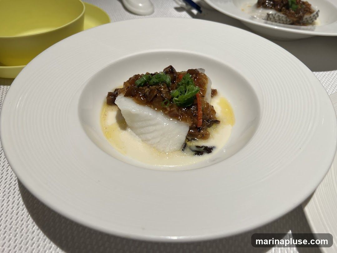A close-up shot of a white plate with a piece of steamed white fish topped with a savory sauce and garnished with green onions and red chili slivers. The fish is served in a shallow white bowl with a textured rim, sitting on a white placemat. A yellow bowl and saucer are visible in the background. Another plate with a similar dish is out of focus in the upper right corner.