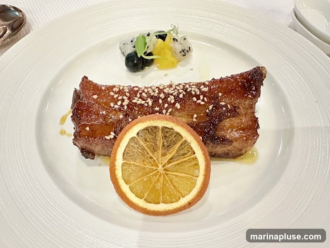 A close-up shot of a gourmet dish featuring a piece of glazed pork belly, sprinkled with sesame seeds. It is garnished with a slice of dried orange and a small portion of fruit salad, including blueberries and dragon fruit. The dish is served on a white, textured plate.