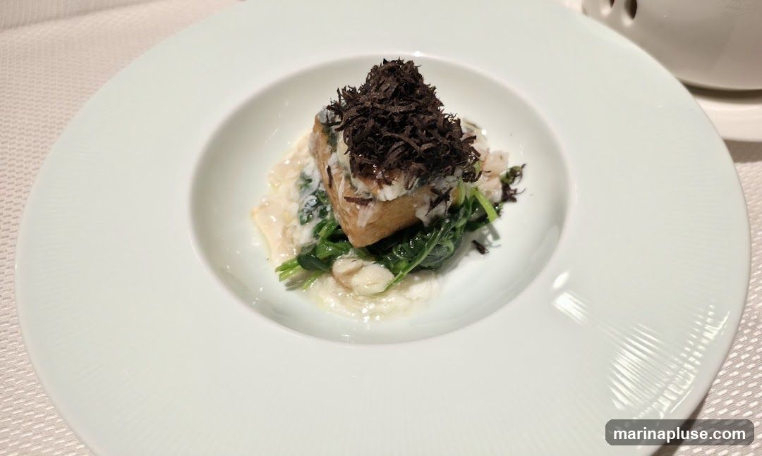 A close-up shot of a finely plated dish featuring a piece of food topped with dark, shredded truffles, served over a bed of wilted greens and a creamy sauce. The dish is presented in a wide, white ceramic bowl with a textured rim, set on a light-colored, textured tablecloth. A portion of a white teapot or similar vessel is visible in the upper right corner.