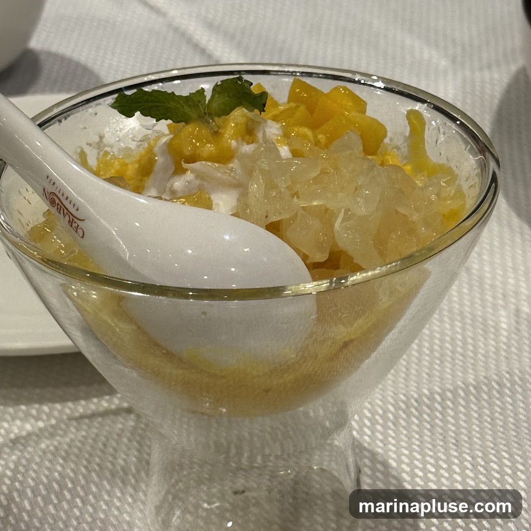 A clear glass bowl filled with a dessert featuring diced mango, pomelo segments, and a creamy white base, possibly ice cream or whipped cream. A white spoon with the logo "CERABON essentials" is placed in the bowl. The dessert is garnished with a sprig of mint. The bowl sits on a white tablecloth.