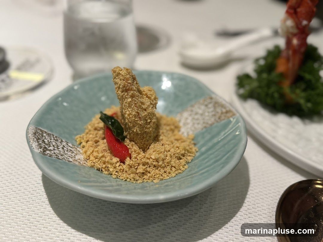 A close-up shot of a small, fried appetizer served on a light blue, textured plate. The appetizer is coated in a crumbly topping and garnished with a red chili pepper and a green leaf. It is presented on a bed of crushed nuts or breadcrumbs. In the background, a glass of water and another dish with a piece of seafood are visible. The table is covered with a white tablecloth.
