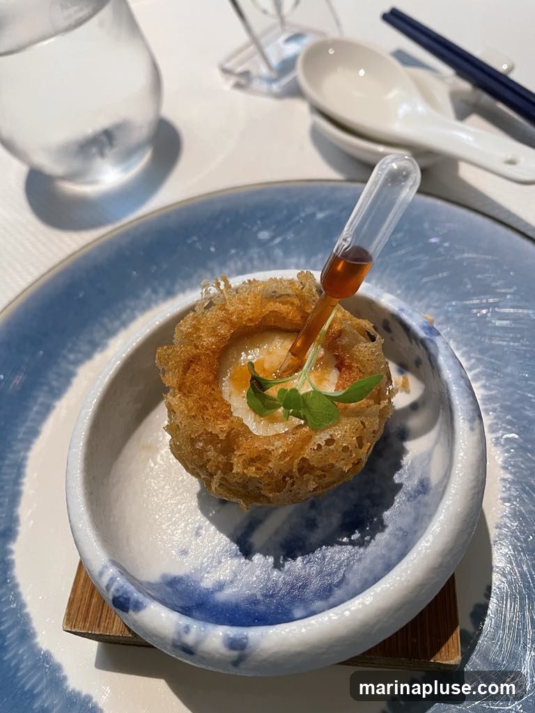 A close-up shot of a delicate, crispy appetizer served in a blue and white ceramic bowl. The appetizer is shaped like a nest and contains a creamy filling, topped with a sprig of green garnish. A small dropper filled with amber liquid is inserted into the appetizer, suggesting a sauce or flavoring to be added. The setting appears to be a restaurant with a glass of water and chopsticks visible in the background.