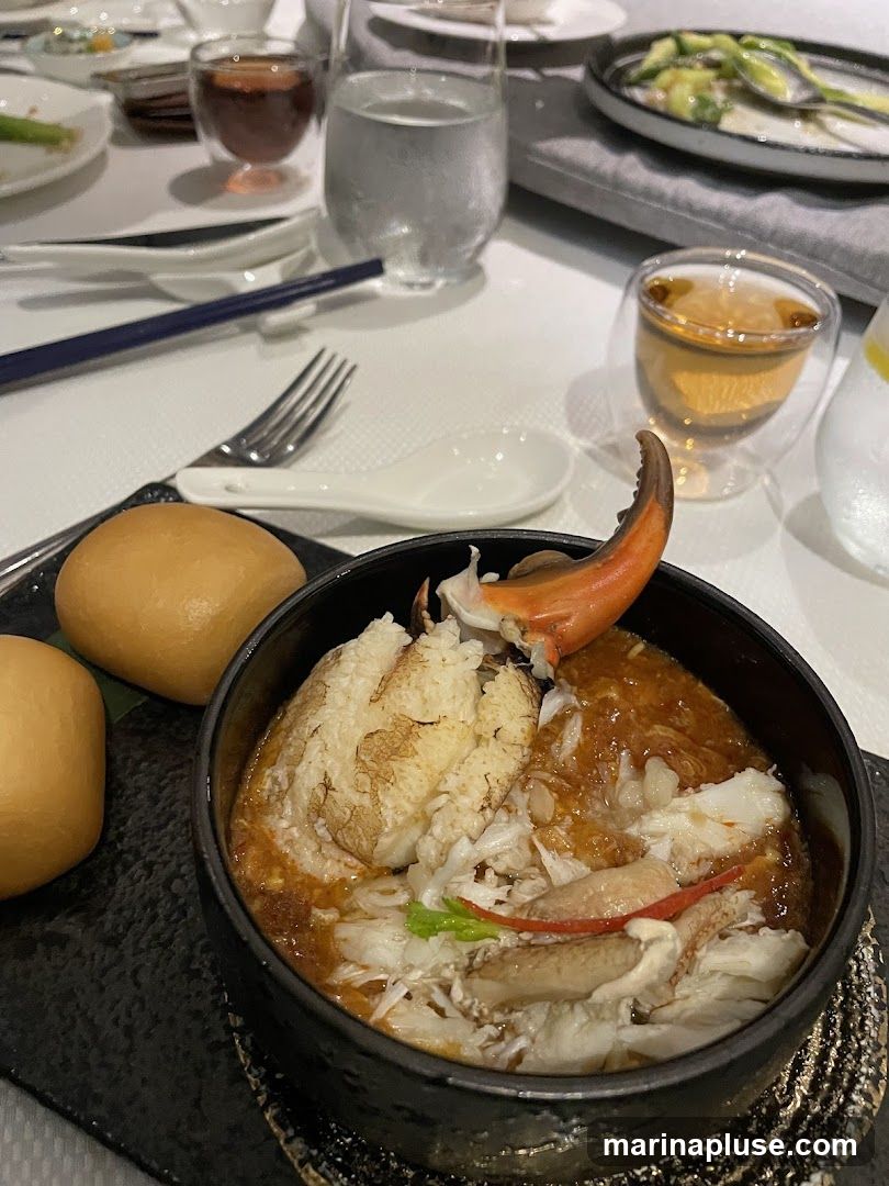 A close-up view of a crab dish served in a black bowl, accompanied by two steamed buns. The crab appears to be shredded and mixed with a savory sauce. The dish is set on a table with other dining elements, including glasses of water and tea, chopsticks, a fork, and spoons.