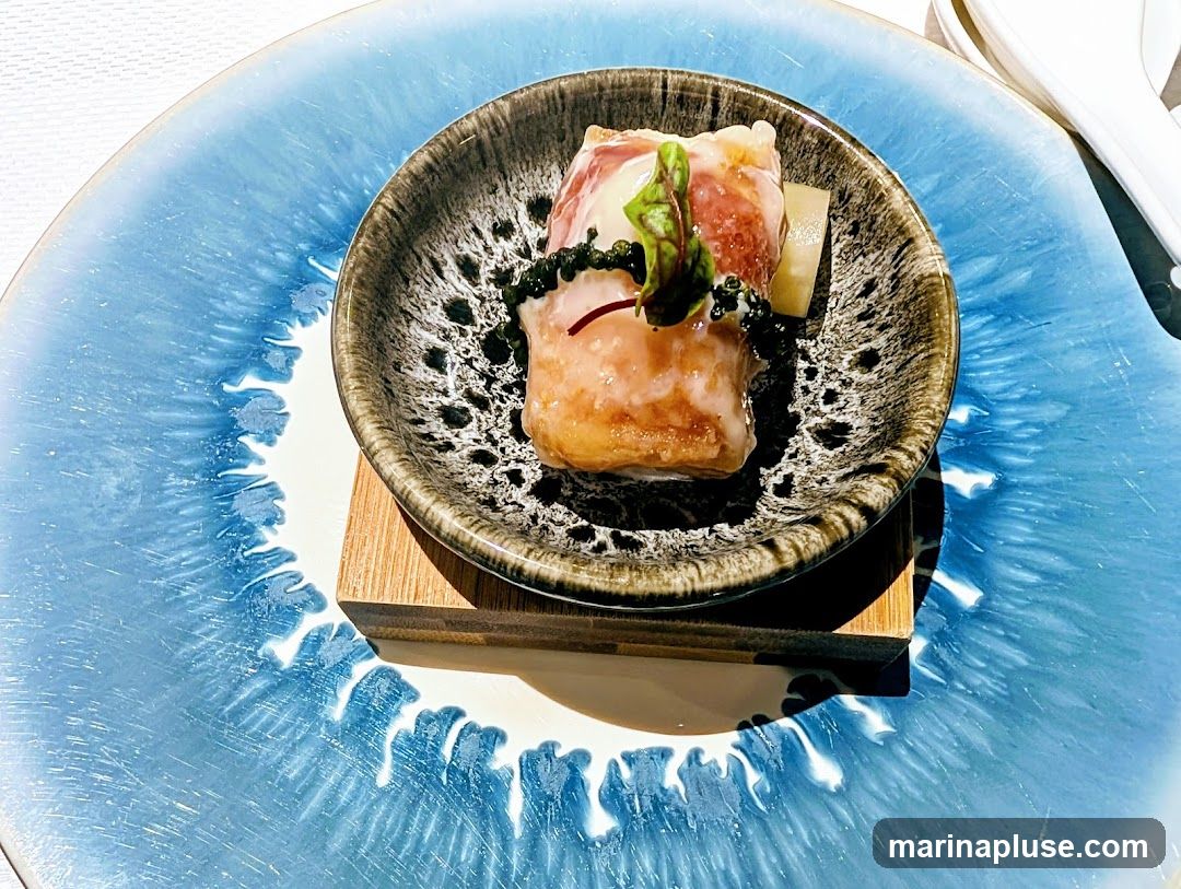 A close-up shot of a single appetizer served in a small, dark, textured bowl. The appetizer appears to be a piece of meat, possibly ham or bacon, wrapped around a filling and topped with a small green leaf and dark beads. It is presented on a larger, light blue plate with a white, wave-like pattern. The dish is placed on a small wooden stand.