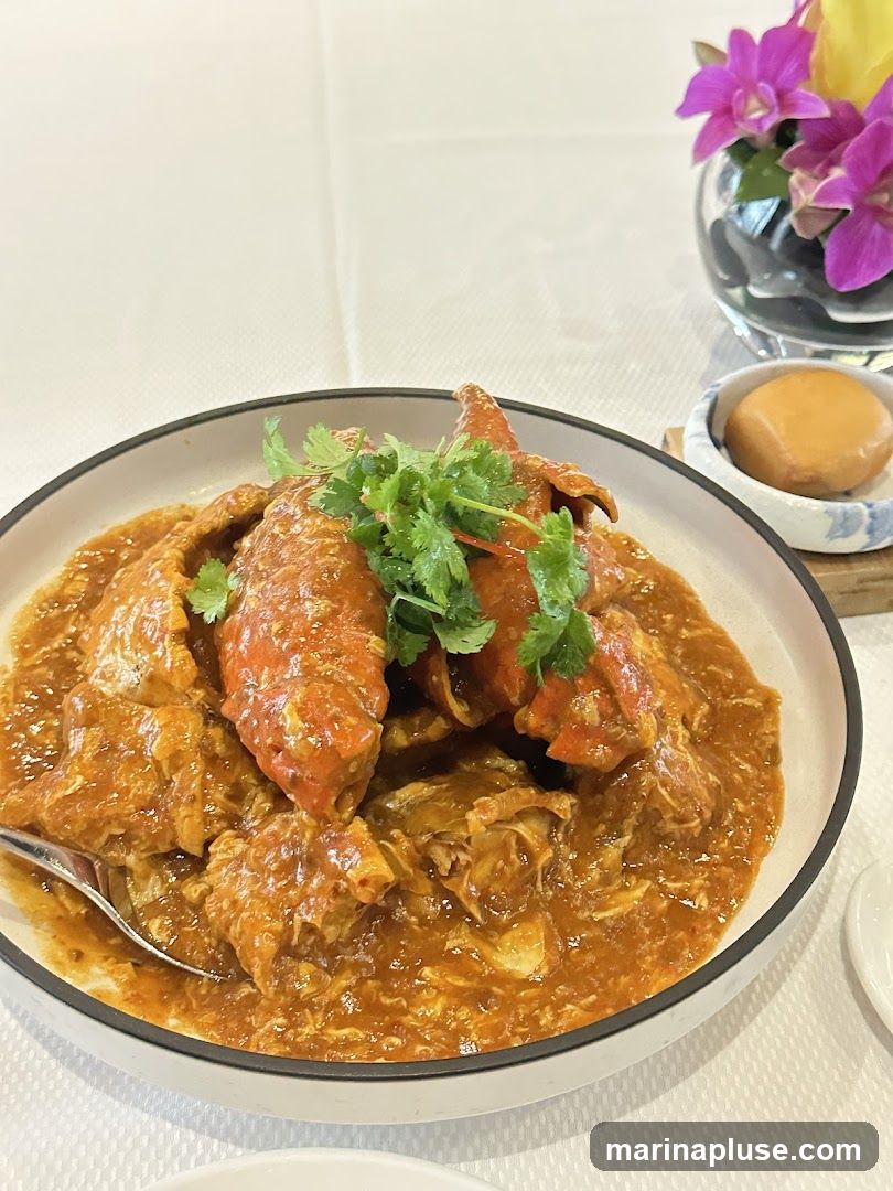A close-up shot of a chili crab dish served in a white bowl with a black rim. The crab pieces are coated in a thick, orange-red sauce and garnished with fresh cilantro. A small side dish with a fried bun is visible to the right, along with a glass vase containing purple orchids and yellow flowers. The table is covered with a white tablecloth.